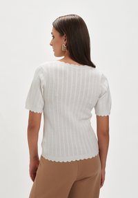 Biały, dzianinowy top z krótkimi rękawami, prążkowaną teksturą, z falbanistym dekoltem i brzegiem, noszony z beżowymi spodniami, widok z tyłu.