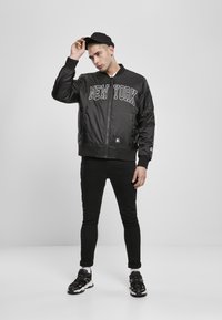 Chaqueta bomber negra con la inscripción "NUEVA YORK", con cierre de cremallera en la parte frontal y puños de canalé. Combinada con jeans negros ajustados y zapatillas negras.