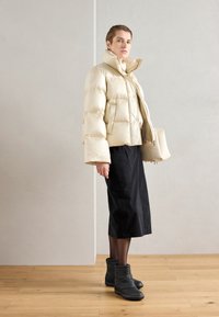 Beige dunjakke med høy krage og oversized ermer. Kombinert med et svart skjørt og ankelhøye svarte støvler. Glatt tekstur, moderne design.