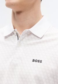 Λευκό πόλο με σχέδιο κυψελωτού πλέγματος, πλάκα με δύο κουμπιά, αντίθετη τρέσα για το γιακά και λογότυπο «BOSS» σε μαύρο στο στήθος.