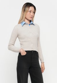 Cárdigan de color beige claro hecho de material suave, con frente de botones, mangas largas y un diseño ajustado, usado sobre una camisa azul de cuello.