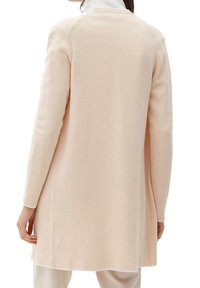 Kvinna som bär en lång, beige stickad cardigan över ljusa byxor, sedd bakifrån mot en vit bakgrund.