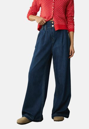 Woman wearing high-waist wide-leg dark blue jeans, red polka-dot buttoned sweater, and beige shoes, hand holding sweater hem.