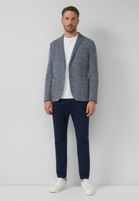 Muž stojaci opretý o bielu stenu, oblečený v modrom saku, bielom tričku, tmavomodrých nohaviciach a bielych teniskách.