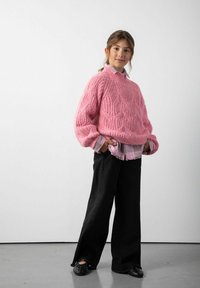 Różowy sweter z dzianiny o teksturowanym wzorze, nałożony na jasnoróżową koszulę z kołnierzykiem. Czarnie szerokie spodnie i czarne buty dopełniają stylizację.