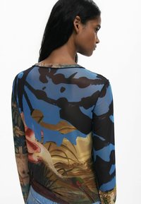 Durchsichtige Langarmbluse mit einem mehrfarbigen Design in Blau-, Schwarz- und Erdtönen, das Vögel und Pflanzen darstellt. Der Saum ist rund.