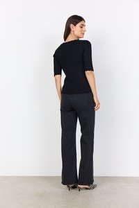 Musta ribbattu neulepaita, jossa on aaltoilevat hihansuut, yhdistettynä mustiin leveälahkeisiin farkkuihin ja raidallisiin korkokenkiin. Siisti tausta.