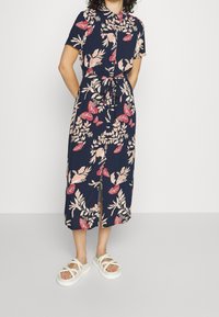 Abito midi con stampa floreale su sfondo blu navy, maniche corte, colletto, vita legata e spacco anteriore. Abbinato a sandali bianchi con cinturino.