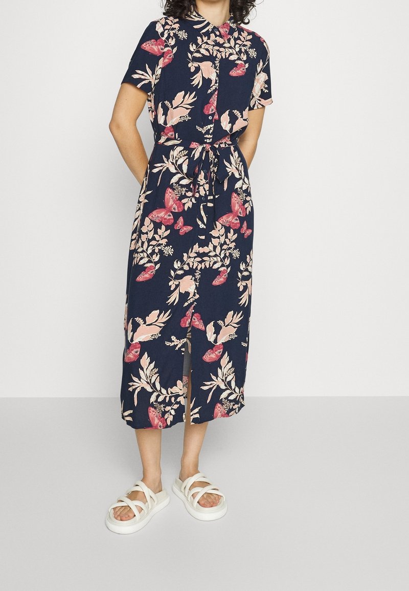 Abito midi con stampa floreale su sfondo blu navy, maniche corte, colletto, vita legata e spacco anteriore. Abbinato a sandali bianchi con cinturino.