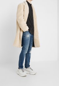 Beżowy puszysty płaszcz na czarnej koszuli, zestawiony z przetartymi niebieskimi dżinsami i białymi trampkami. Płaszcz ma klapowy kołnierz i guziki z przodu.