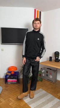 Melna Adidas jakna ar rāvējslēdzēju un baltām sloksnēm, melna ādas bikses un brūni zābaki. Iekštelpu vide ar krāsainu dekoru un mēbelēm.