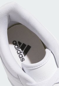 White sneaker with textured fabric and smooth accents. Features black "adidas" logo on the insole, rounded collar, and stitched details.