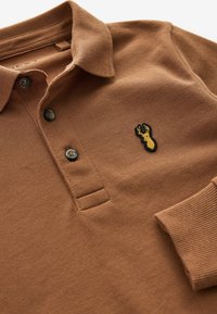 Polo en coton marron avec une texture tissée, trois boutons et un logo de homard jaune brodé sur le côté gauche de la poitrine.