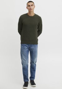 Grüner strukturierter Strickpullover mit rundem Ausschnitt, kombiniert mit hellblauen Jeans und schwarzen Sneakers. Schlichtes Design ohne sichtbare Muster.
