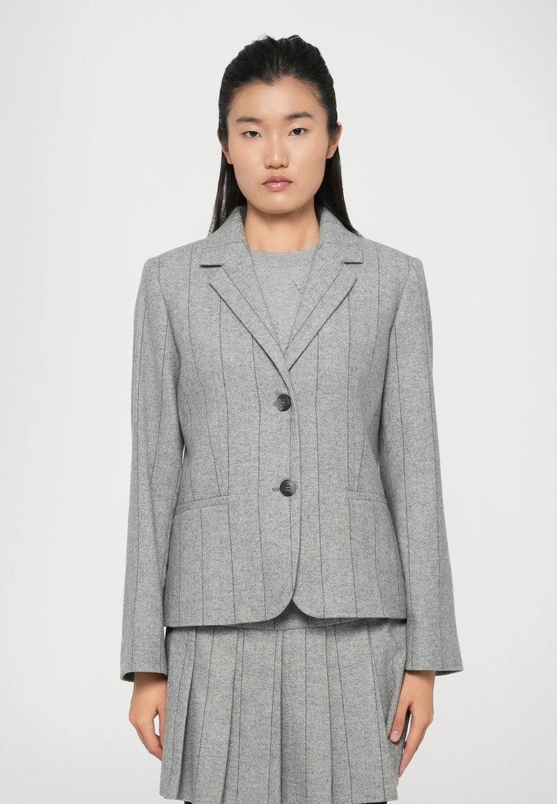 Mulher a usar um blazer cinza ajustado com costuras verticais e botões, combinado com uma saia plissada correspondente e um suéter.