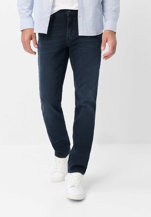 Uomo che indossa jeans blu scuro, sneaker bianche e una camicia a righe azzurra parzialmente infilata, in piedi su uno sfondo bianco uniforme.