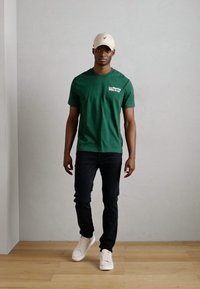 Grønn bomulls t-skjorte med hvit tekst, kombinert med svarte denim jeans og hvite sneakers. Beigefarget caps fullfører den avslappede stilen.