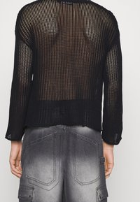 Schwarzer Strickpullover mit lockerem Design und offenem Muster, mit langen Ärmeln und einem cropped Rückenteil. Kombiniert mit grauen Denimbomberhosen.