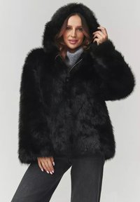 Czarna kurtka z sztucznego futra z kapturem o puszystej teksturze, z przodem zapinanym na guziki i szerokimi rękawami, noszona na szarym swetrze i ciemnych dżinsach.