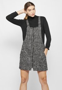 Črno-bela tweed obleka s širokimi naramnicami, sprednjimi žepi in teksturiranim vzorcem, preplastena s črnim puloverjem s turtleneck ovratnikom.