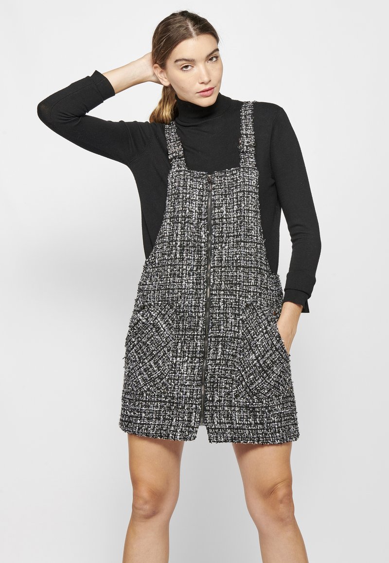 Črno-bela tweed obleka s širokimi naramnicami, sprednjimi žepi in teksturiranim vzorcem, preplastena s črnim puloverjem s turtleneck ovratnikom.