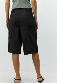 Schwarze Baumwoll-Bermuda-Shorts erreichen die Mitte der Waden, haben einen geraden Schnitt, Seitentaschen und einen umgeschlagenen Saum. Glatte Textur, lässiges Design.