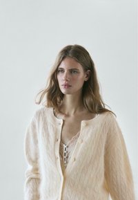 Pale pink knitted cardigan with a cable pattern, round neckline, and button front. Layered over a lace camisole, showcasing texture contrast.