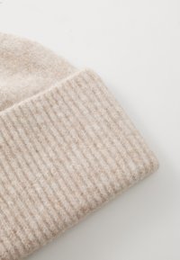Beżowa wełniana czapka z ściągaczem, o miękkim, textured wykończeniu. Materiał wygląda na ciepły i przytulny, odpowiedni na zimną pogodę.