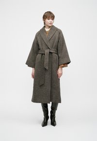 Woman wearing a long belted gray textured coat over a brown sweater and black knee-high boots, standing against a plain light background.