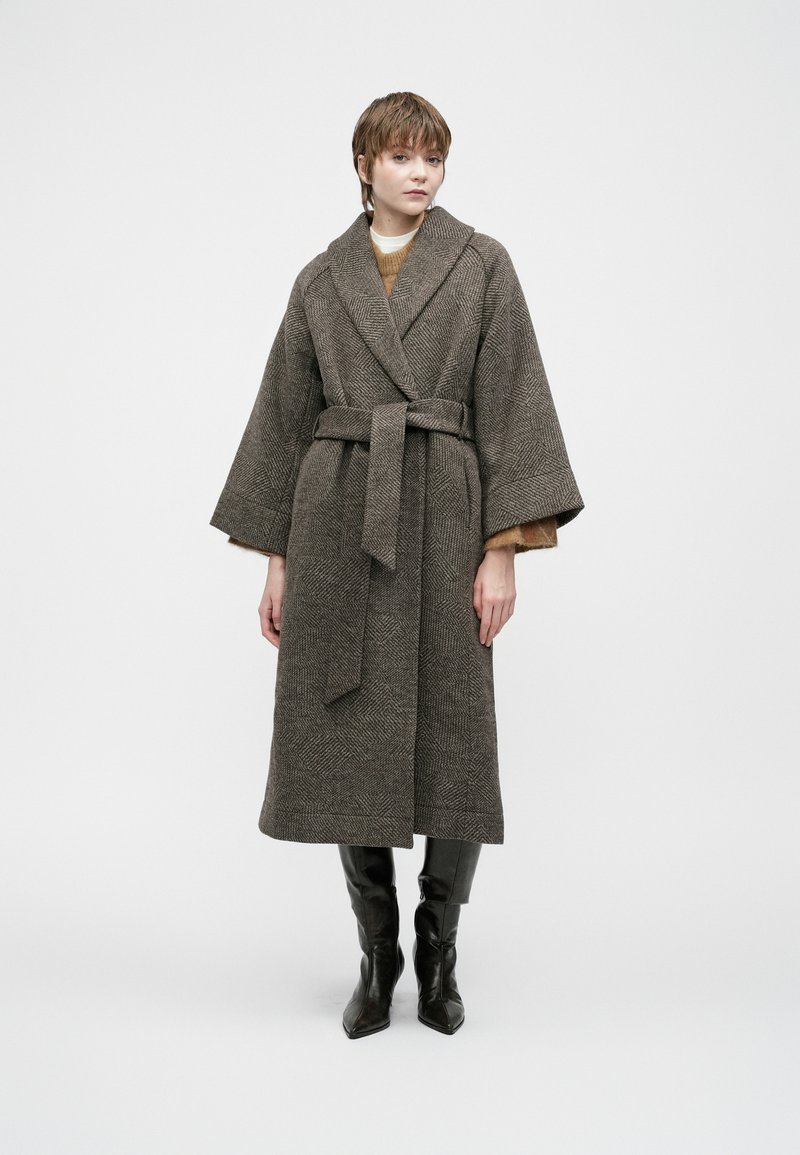 Woman wearing a long belted gray textured coat over a brown sweater and black knee-high boots, standing against a plain light background.