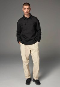Sort langærmet polo med to brystlommer og knapper, parret med beige bukser og sorte sneakers, sat mod en neutral baggrund.