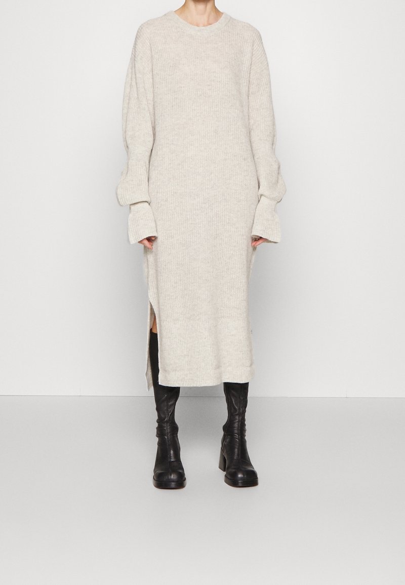 Personne portant une longue robe pull en tricot beige avec des manches surdimensionnées et des bottes noires à talons montant jusqu'aux genoux, debout devant un fond uni.