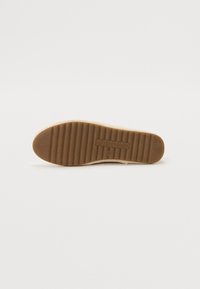 Gummisåle med en rillestruktur, i en lys tan-farge og preget logo. Kanten er sydd med et mykt beige tekstil.