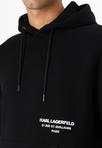 Čierna mikina s kapucňou, ktorá má klokanie vrecko, šnúrky a biely textový logo s detailmi adresy. Mäkký materiál, ležérny dizajn a štandardný strih.