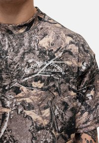 Camo-patterned t-shirt in earthy tones of brown and green, with a round neckline and text logo in white: “GOOD FOR NOTHING BRITISH ATELIER EST 2011.”