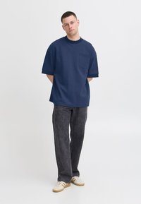 Tumed tooriga ülisuure T-särk lühikeste varrukatega, millel on rinnatasku ja ribiga krae, kombineeritud tumedate teksade ja valgete tossudega.