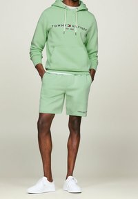 Felpa verde chiaro con tasca frontale e logo, abbinata a pantaloni corti coordinati. Sneakers bianche completano l'outfit. Tessuto morbido e vestibilità rilassata.