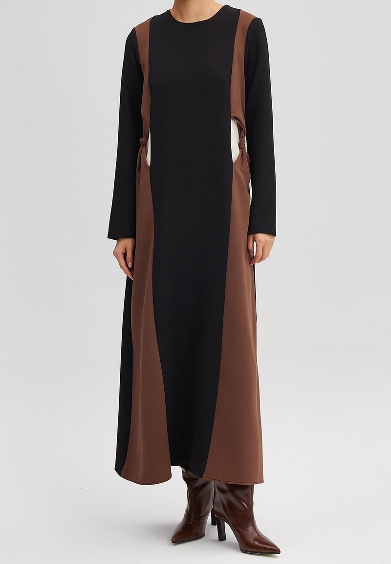 Robe à manches longues présentant des panneaux noirs, marron et crème. Le tissu semble lisse avec des découpes latérales et un détail noué. Portée avec des bottes à talons marron.
