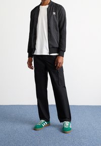 Svart bomberjacka med texturerade paneler, vit logotyp, kombinerad med en vit t-shirt, svarta cargobyxor och gröna sneakers med vita ränder.