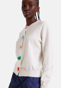 Cremefarbener Cardigan mit rundem Ausschnitt, versehen mit mehrfarbigen, herzförmigen Knöpfen. Weiche Textur und figurbetontes Design mit geripptem Saum.