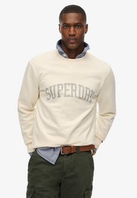 Felpa color crema con la scritta "SUPERDRY" in grigio chiaro sulla parte anteriore, indossata sopra una camicia blu con colletto, abbinata a pantaloni verde oliva.