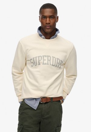 Crème-kleurig sweatshirt met "SUPERDRY" in lichtgrijs over de voorkant, gelaagd over een blauwe blouse, gepaard met olijfgroene broek.