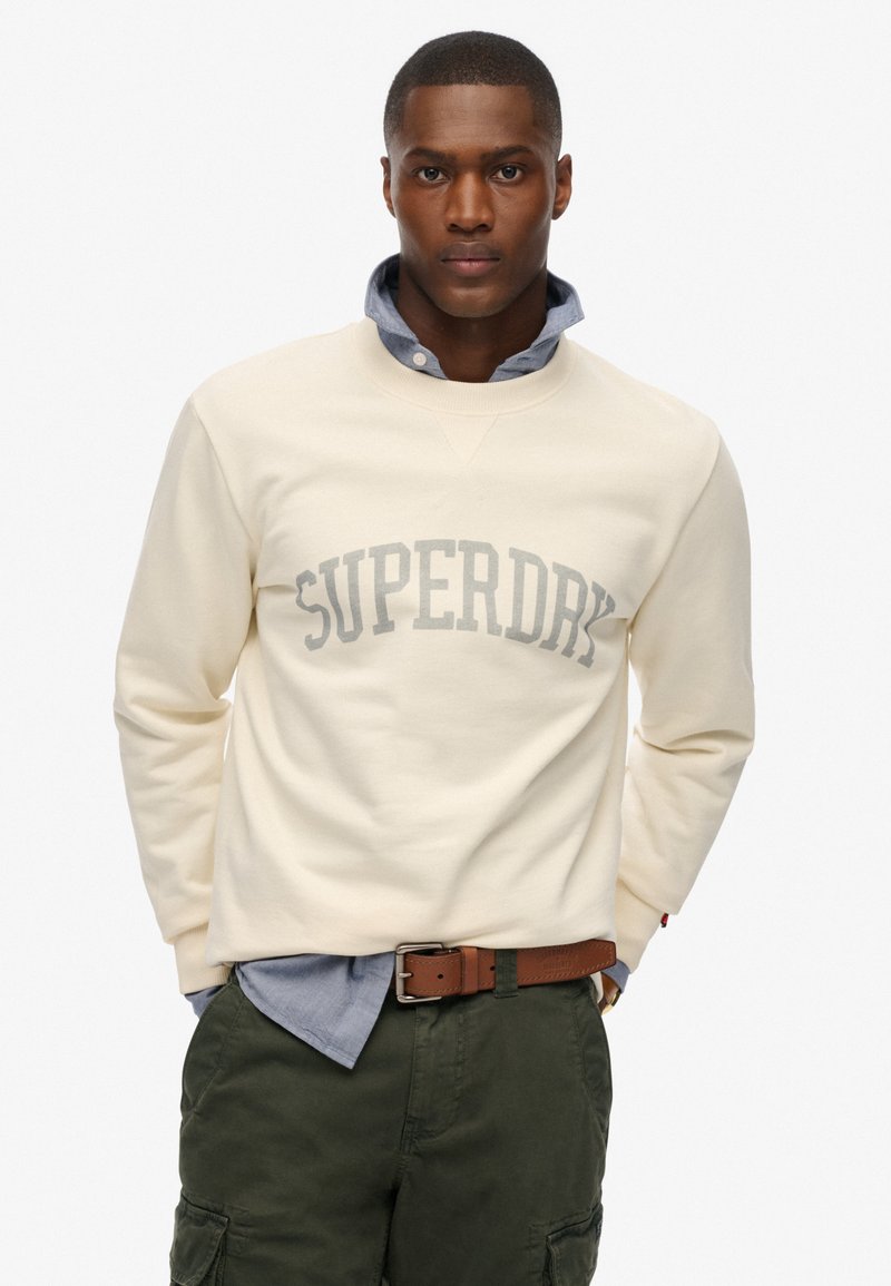 Felpa color crema con la scritta "SUPERDRY" in grigio chiaro sulla parte anteriore, indossata sopra una camicia blu con colletto, abbinata a pantaloni verde oliva.