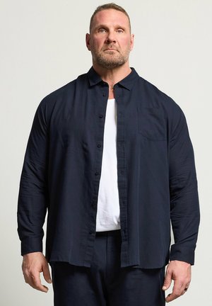 Mand med kort hår og skæg iført en mørk marineblå skjorte med knapper over en hvid T-shirt, stående mod en ensartet lys baggrund.