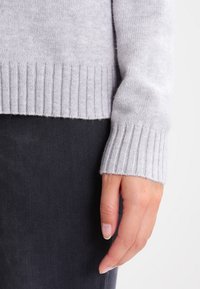 Lichtgrijze gebreide trui met ribbels aan de manchetten en onderkant, gemaakt van zacht materiaal. Zichtbare hand die naast donkere broek ligt.