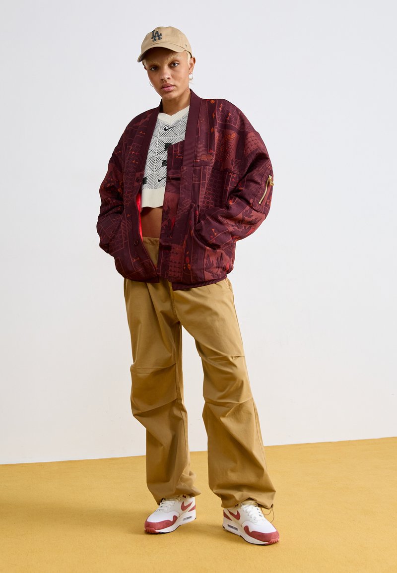 Bordeaux gemusterte übergroße Jacke mit kontrastierendem Innenfutter, beigefarbener Cropped-Top, khakifarbenen weiten Hosen und weißen Sneakers mit roten Akzenten.