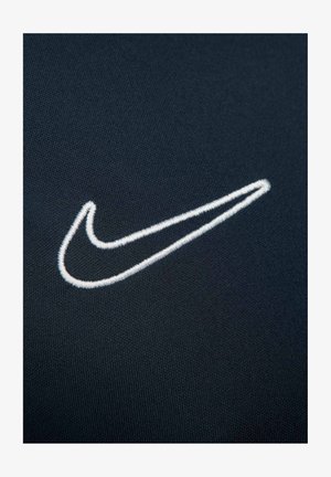 Nike-logo in witte borduursel op een gestructureerde donkere stof achtergrond, met een slanke, gebogen vorm en een onderscheidende uitstraling.