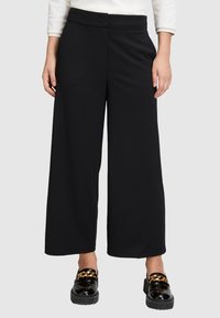 Frau in schwarzen, weit geschnittenen, cropped Hosen und glänzenden schwarzen Slippern mit goldener Kettendetail, vor einem schlichten Hintergrund stehend.