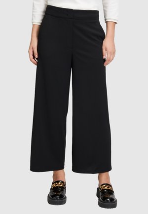 Femme portant un pantalon large cropped noir et des mocassins noirs brillants avec détail en chaîne dorée, se tenant devant un fond uni.