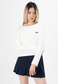 Mladá žena se rovnými ombre vlasy, oblečená v bílé haleně s dlouhými rukávy od značky GAP a námořnické plisované sukni, stojící se zkříženýma rukama.